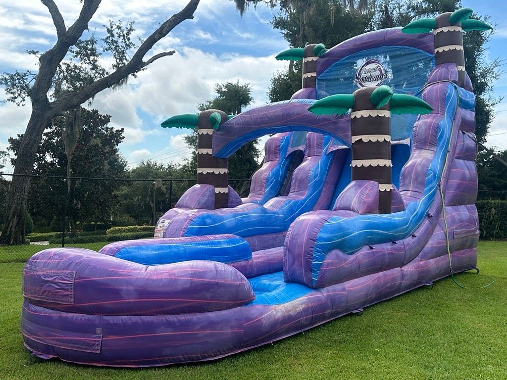 A purple and blue inflatable water slide with palm tree decorations set on a green lawn.