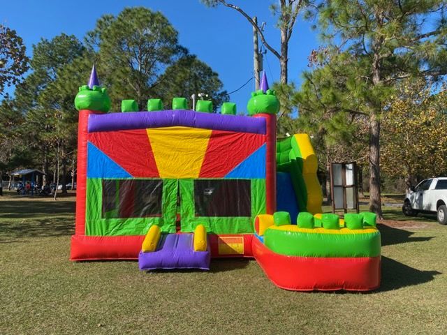 A colorful, multi-section inflatable bounce house and slide set up on a grassy lawn surrounded by trees.