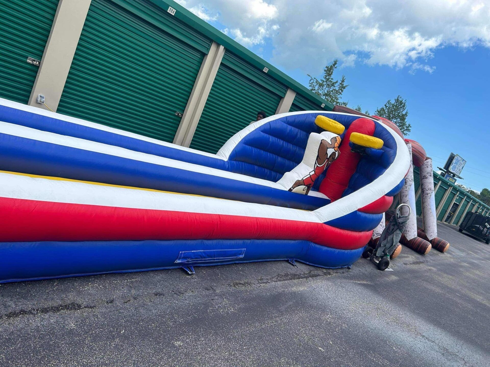 A long, inflatable blue, white, and red obstacle course set up in an outdoor parking lot next to storage units.