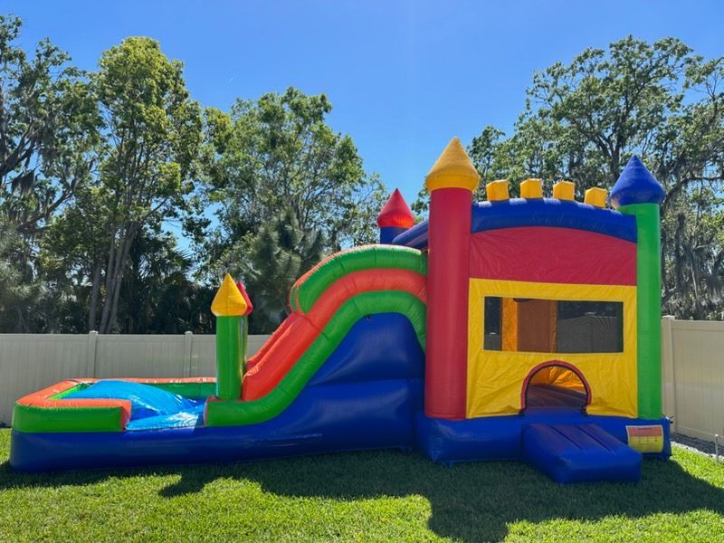 A multi-colored inflatable bounce house with a slide and a pool attached, set on a grassy lawn in front of a white fence.