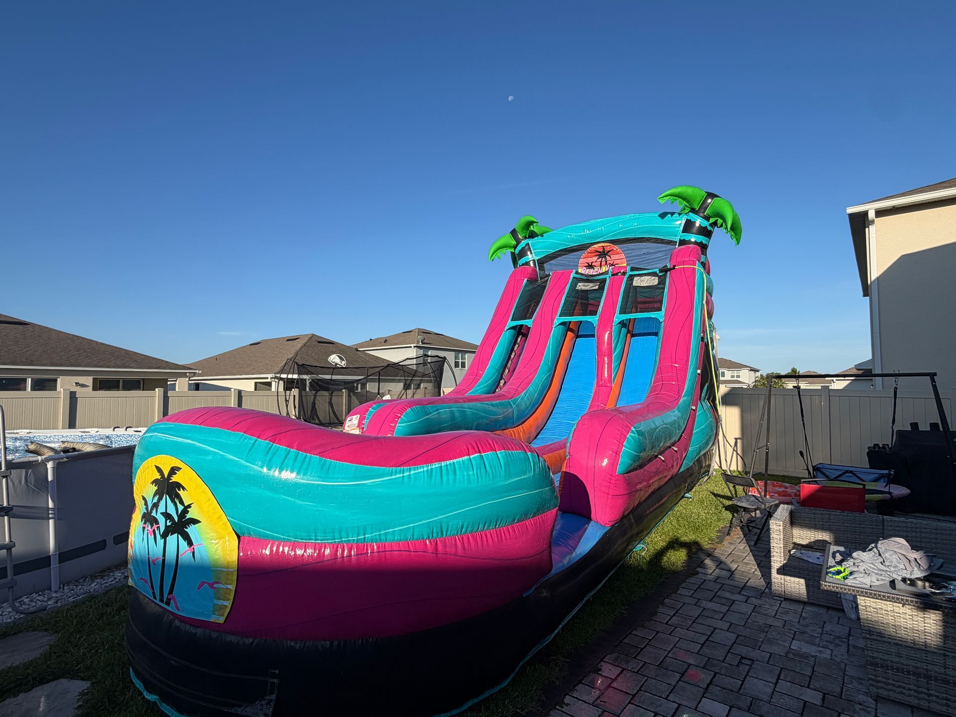 Bright pink and turquoise inflatable water slide with palm trees in a backyard under a clear blue sky