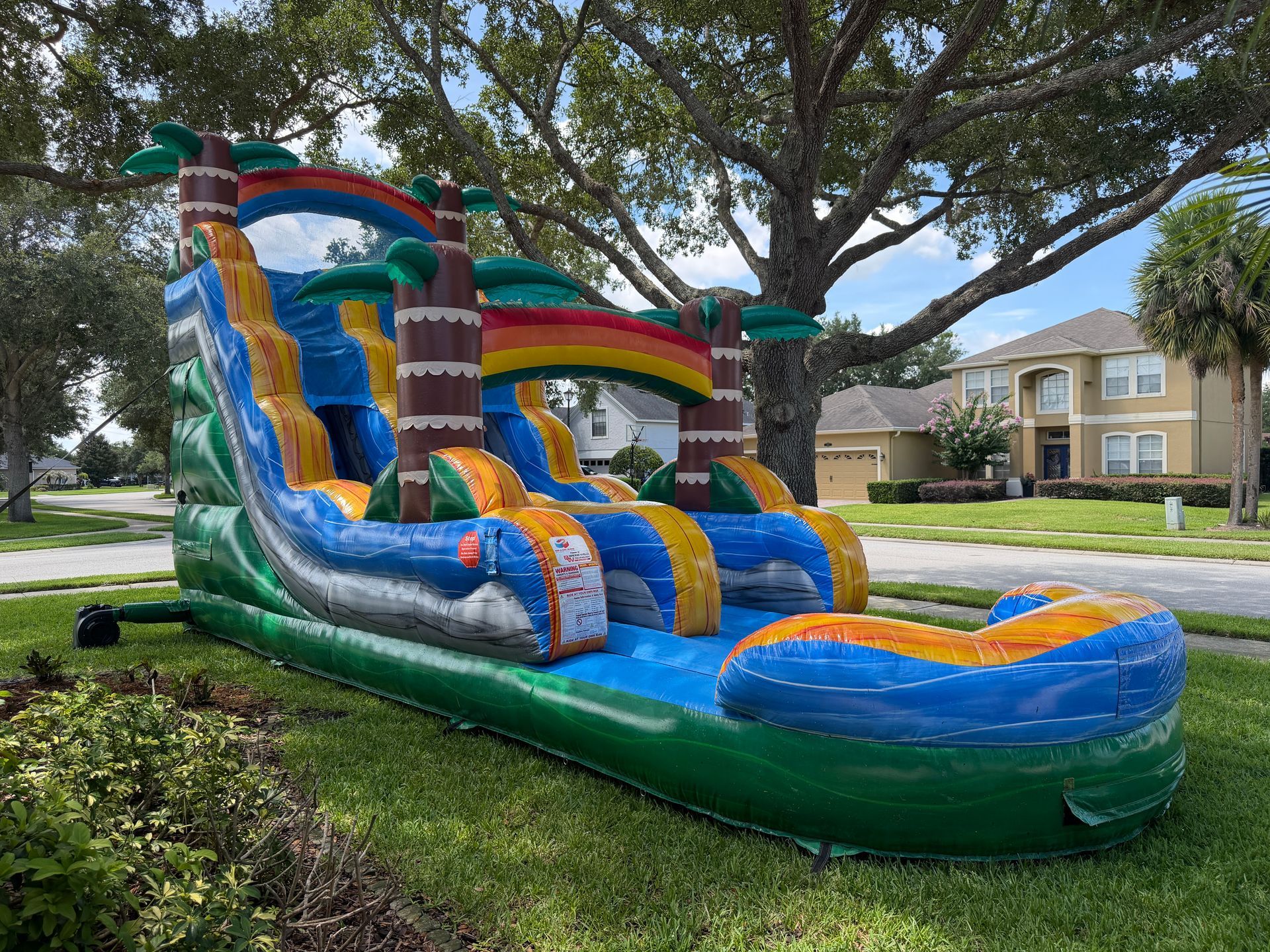 Colorful inflatable backyard water slide on a lawn under trees, with houses in the background