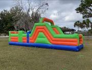 A long, inflatable obstacle course with green, orange, and blue panels, set on a grassy field under a cloudy sky.