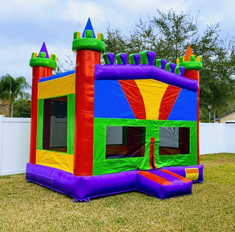 A colorful, multi-towered bouncy castle for children sits on a grassy lawn in front of a white privacy fence.