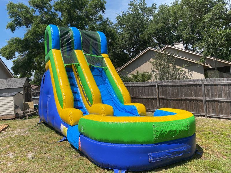 A blue, green, and yellow inflatable water slide with a splash pool, set in a grassy backyard near a wooden fence.