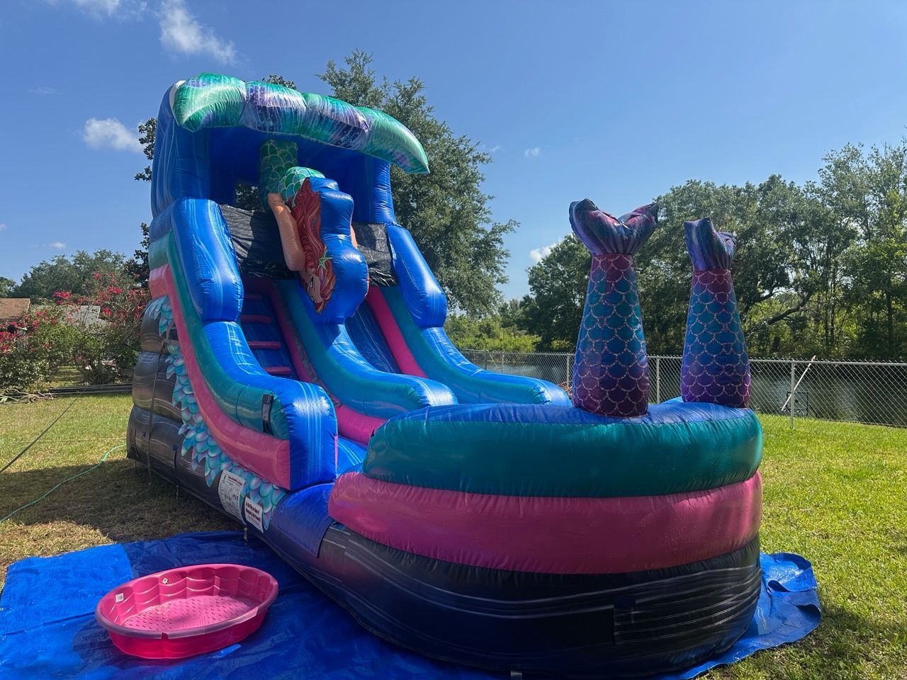 A brightly colored inflatable mermaid-themed water slide stands on a lawn with a small pink pool at the base.