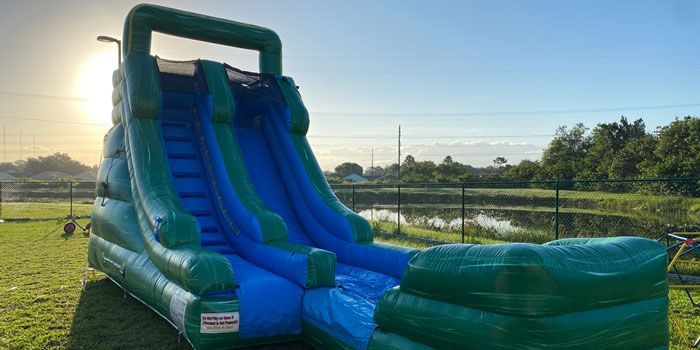 A tall, green and blue inflatable water slide sits outdoors on a grassy lawn near a pond under a bright, sunny sky.