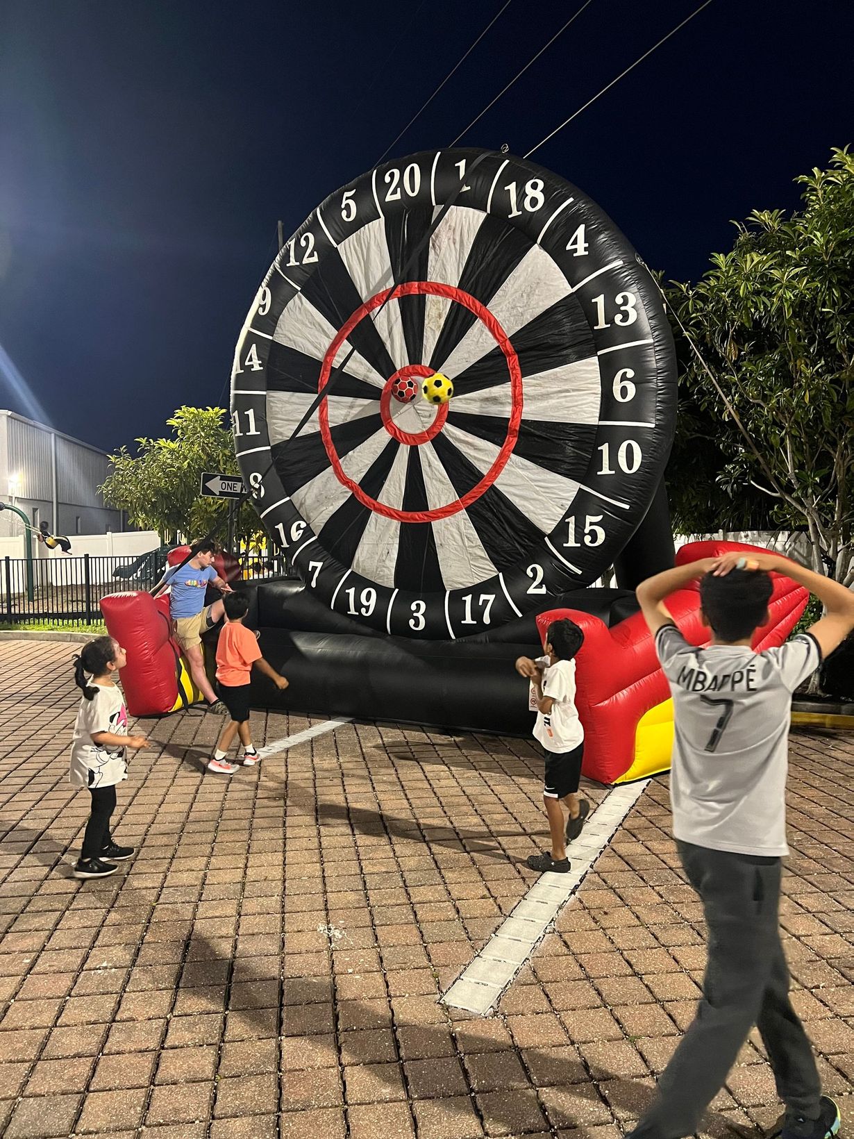 Children play a giant inflatable velcro dartboard game outdoors at night on a paved surface.
