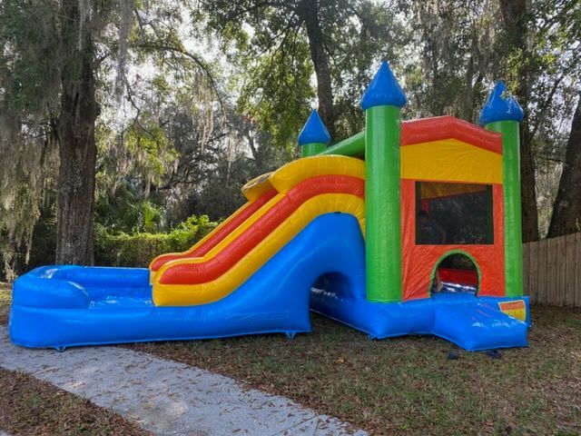 A bright, colorful inflatable bounce house with a slide and a pool attached, set on a grassy lawn under trees.