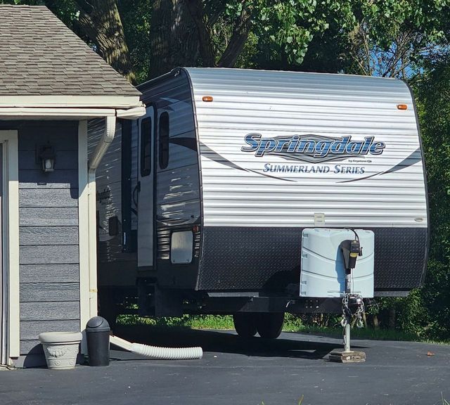 White and gray Spree travel trailer parked beside a house with a white utility hookup and black driveway.