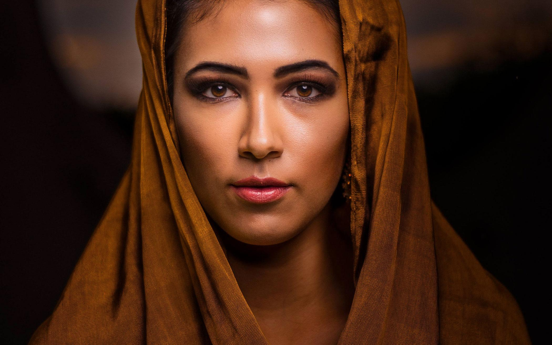 Close-up portrait of a person in a brown hooded scarf against a dark background
