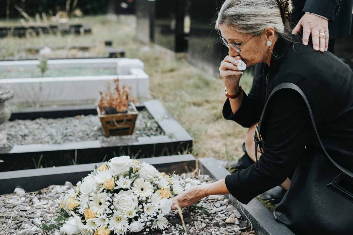 Woman at a cemetery, wiping tears, placing flowers on a grave. Another person's hand on her shoulder.