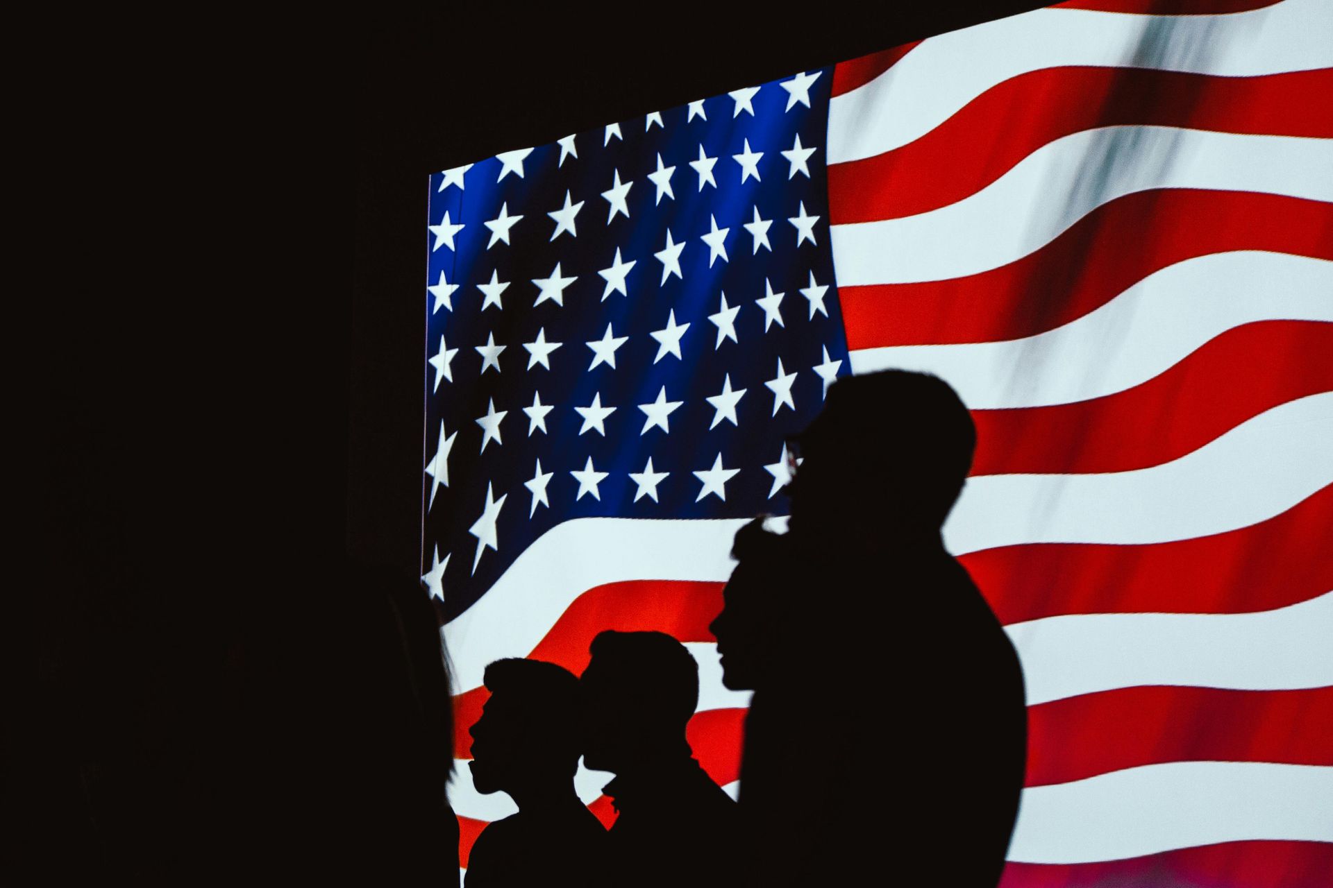 Silhouetted figures stand before a waving American flag.