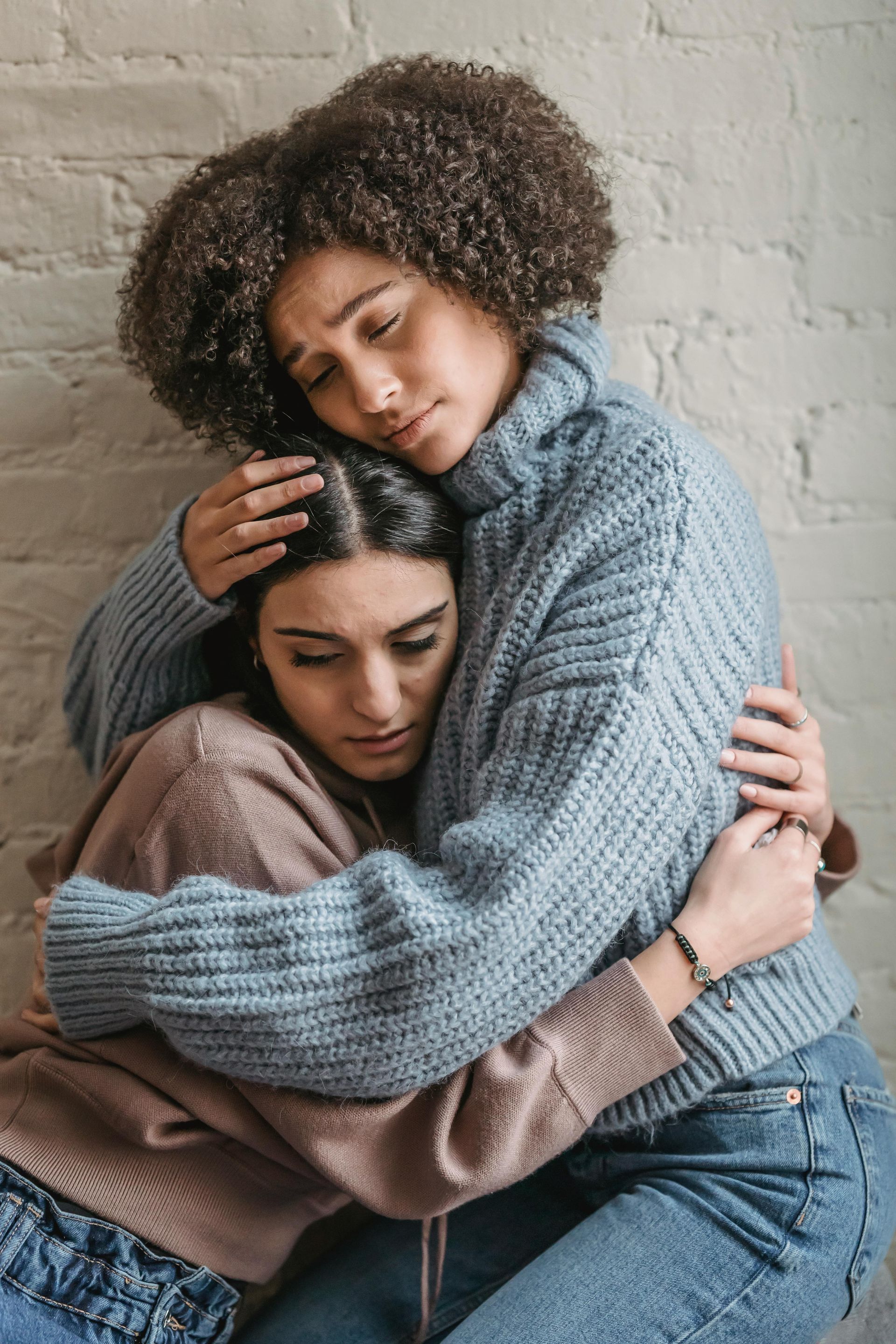 Two people embracing, one comforting the other. Both have sorrowful expressions; against a white brick wall.
