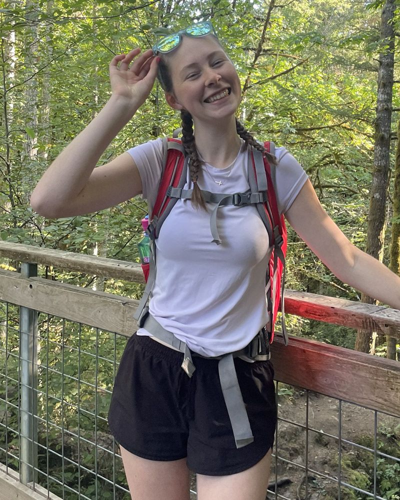 Woman on a wooden bridge in a forest, wearing hiking gear and smiling. Sunglasses on her head.