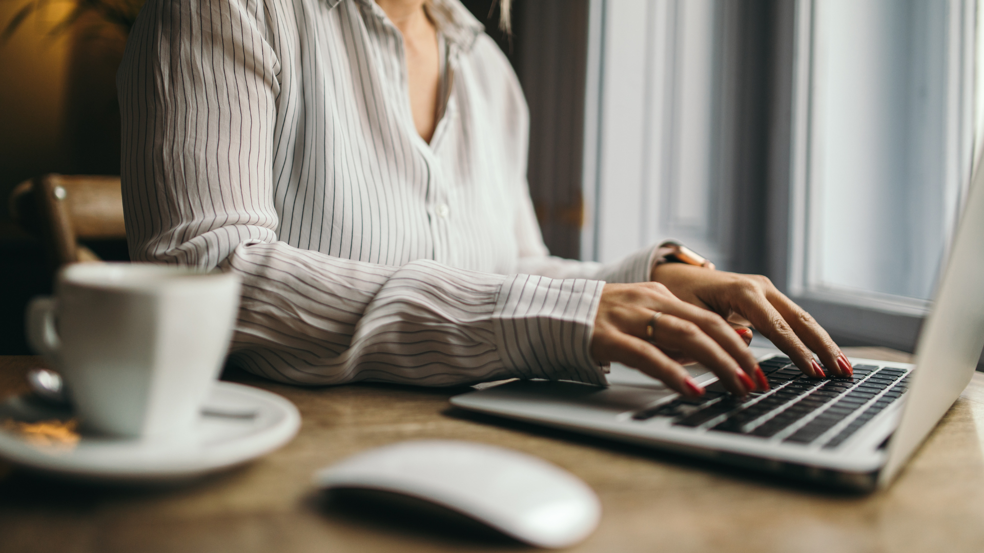 Woman typing on a laptop at a table by a window, coffee and mouse in view.