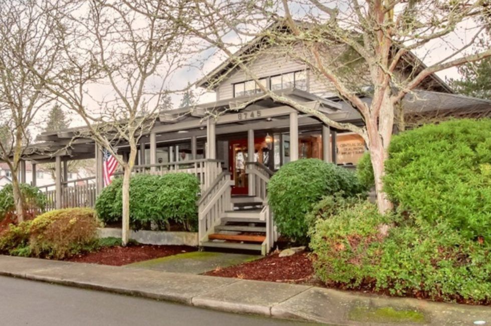 Rustic building with porch, steps, and American flag, framed by trees and bushes.