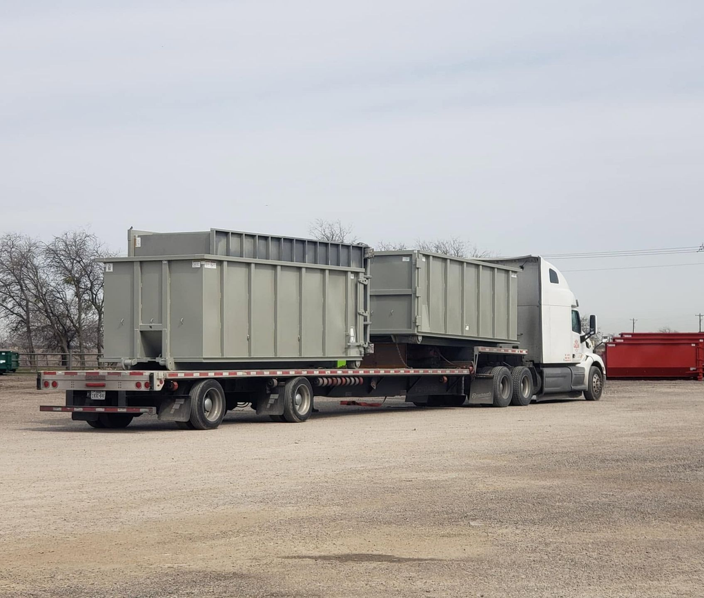 White semi-truck hauling two large gray industrial containers on a flatbed in a gravel lot.
