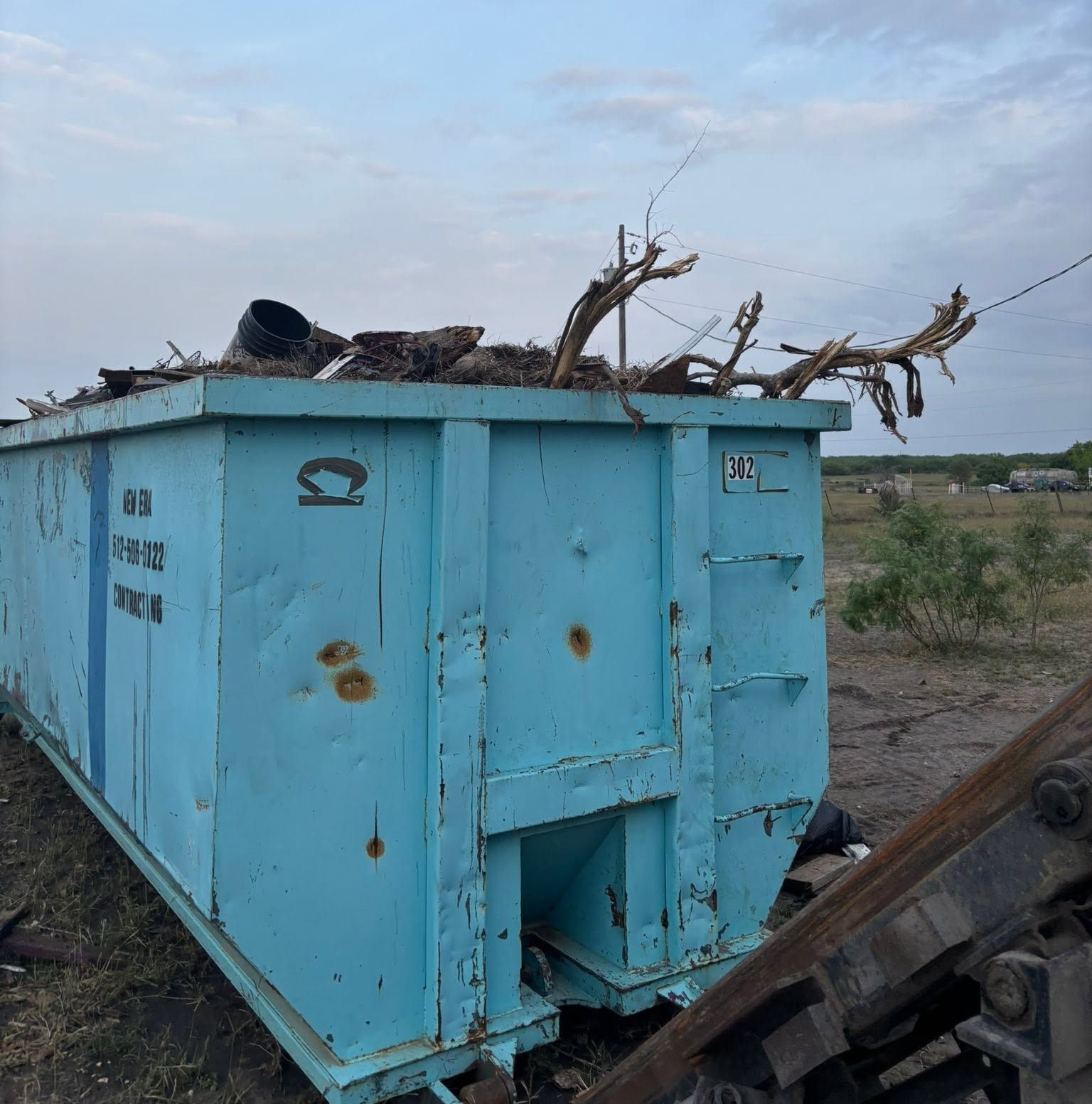 Light blue metal dumpster outdoors on a dirt lot, with debris piled on top.