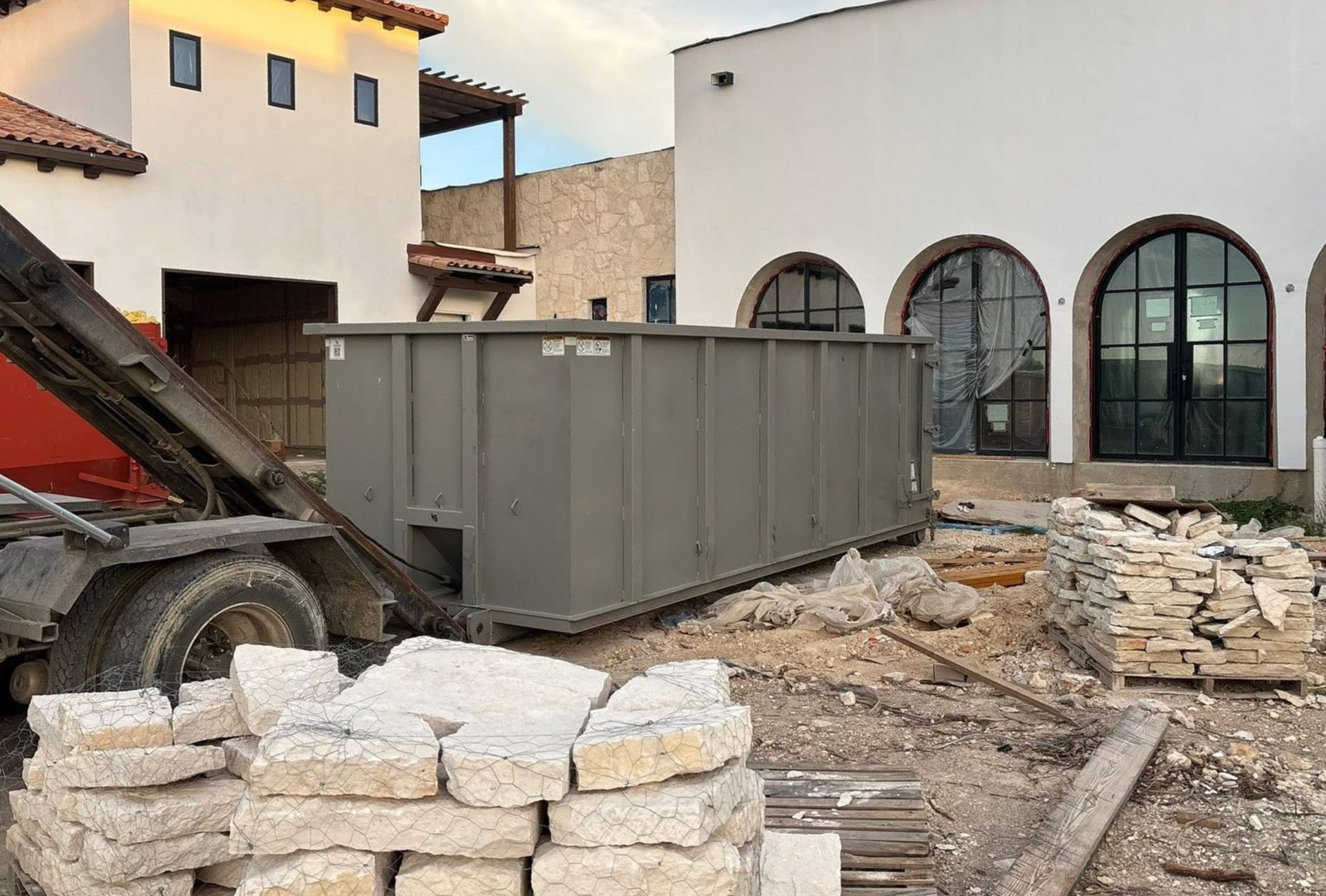 Construction site with large gray dumpster, rubble, sandbags, and a red dump truck beside a white building