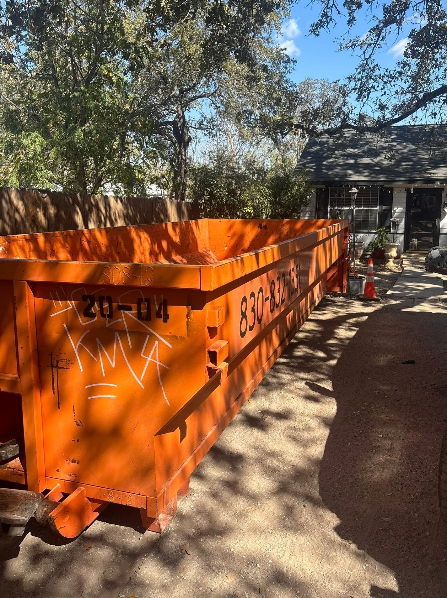 Orange metal dumpster in a shaded yard near a house, with trees and a dirt path nearby.