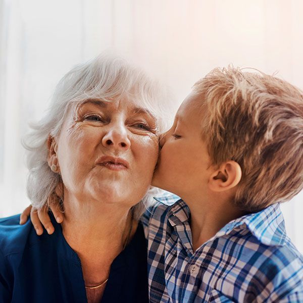 Older woman smiling as a child kisses her cheek, both in blue plaid shirts on a soft indoor background