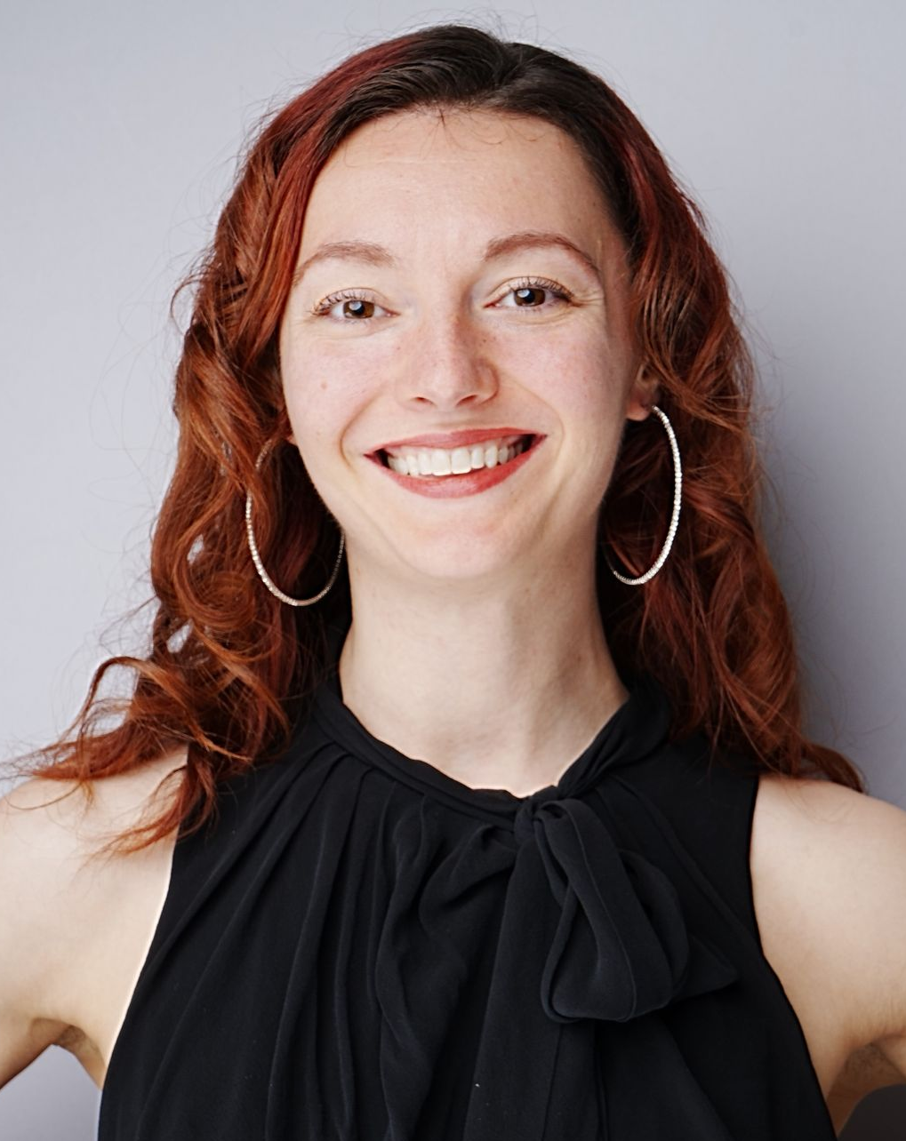 Smiling person with red hair and hoop earrings in a black sleeveless top on a gray background