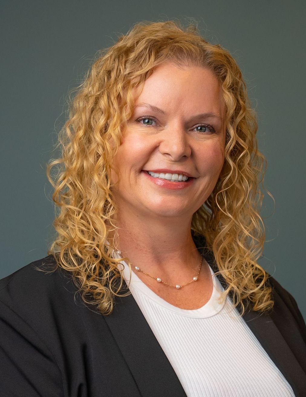 Phobe Todorof Woman with curly blonde hair, smiling, wearing a black blazer and white top, against a dark blue background.