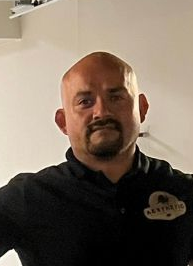 Rene Holguin Man in a black polo shirt standing indoors against a light wall, smiling at the camera.