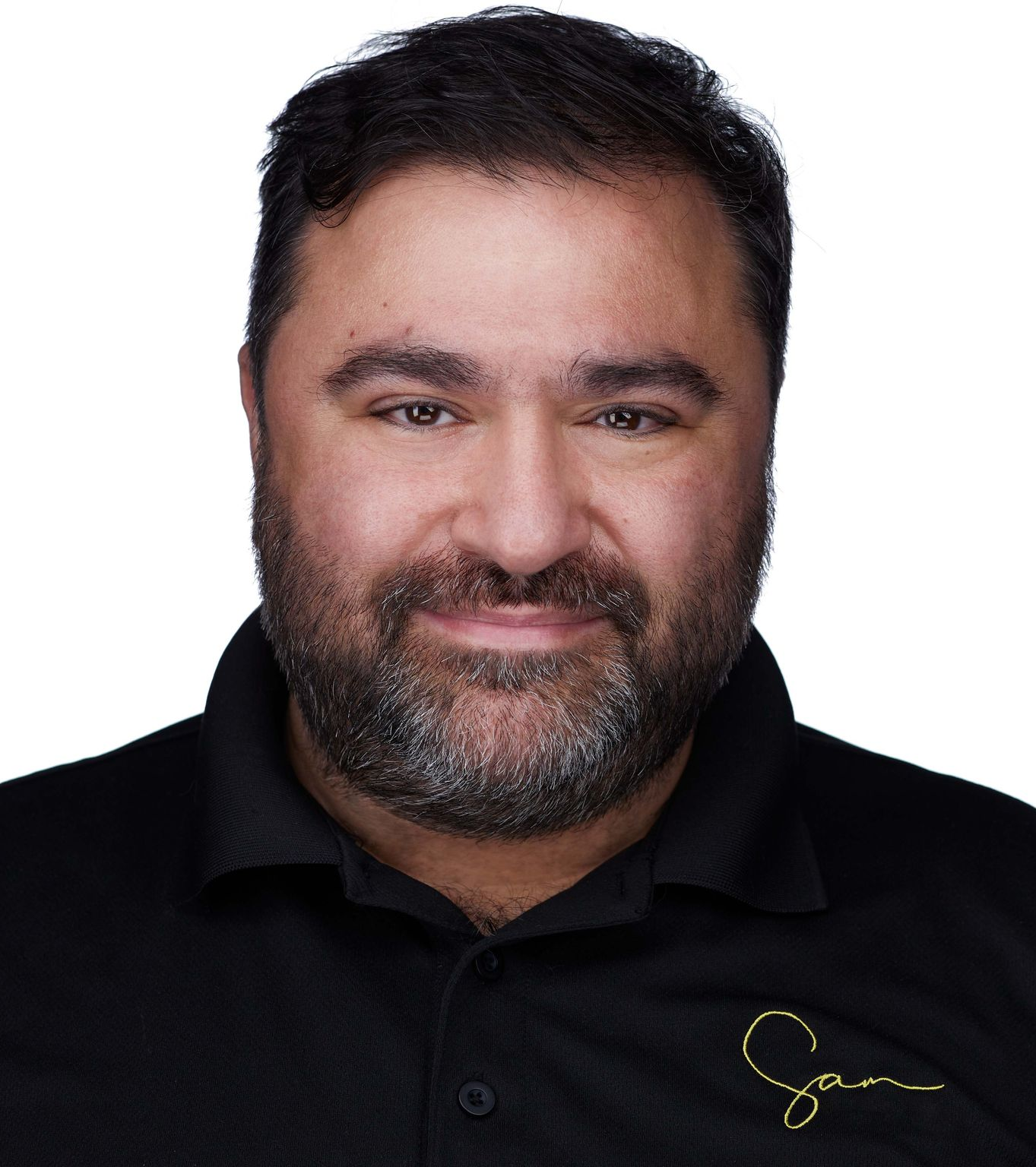 Man with dark hair and beard smiling, wearing a black shirt with a gold logo.