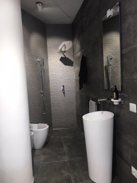 Bagno moderno con pareti rivestite di piastrelle grigie, sanitari bianchi, specchio e un alto lavabo a colonna bianco.