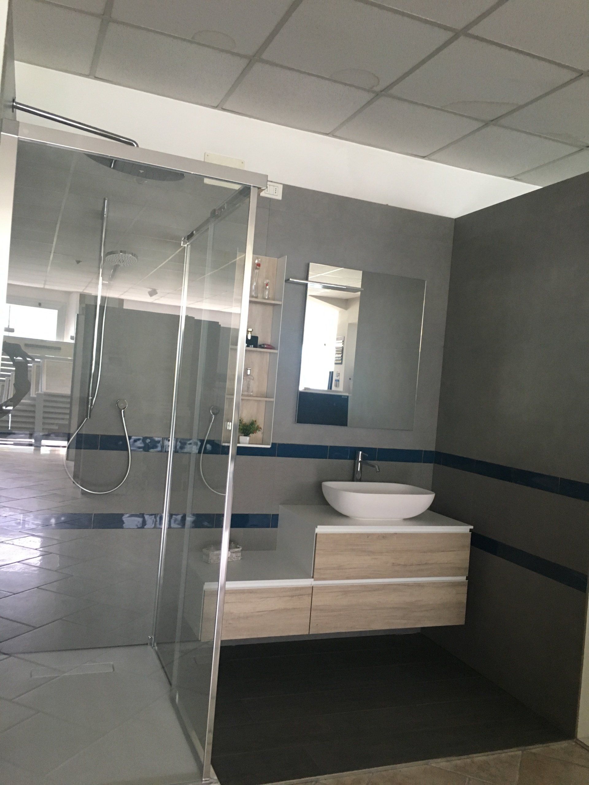 Bagno moderno con box doccia in vetro e lavabo a parete sopra un mobiletto in legno chiaro.