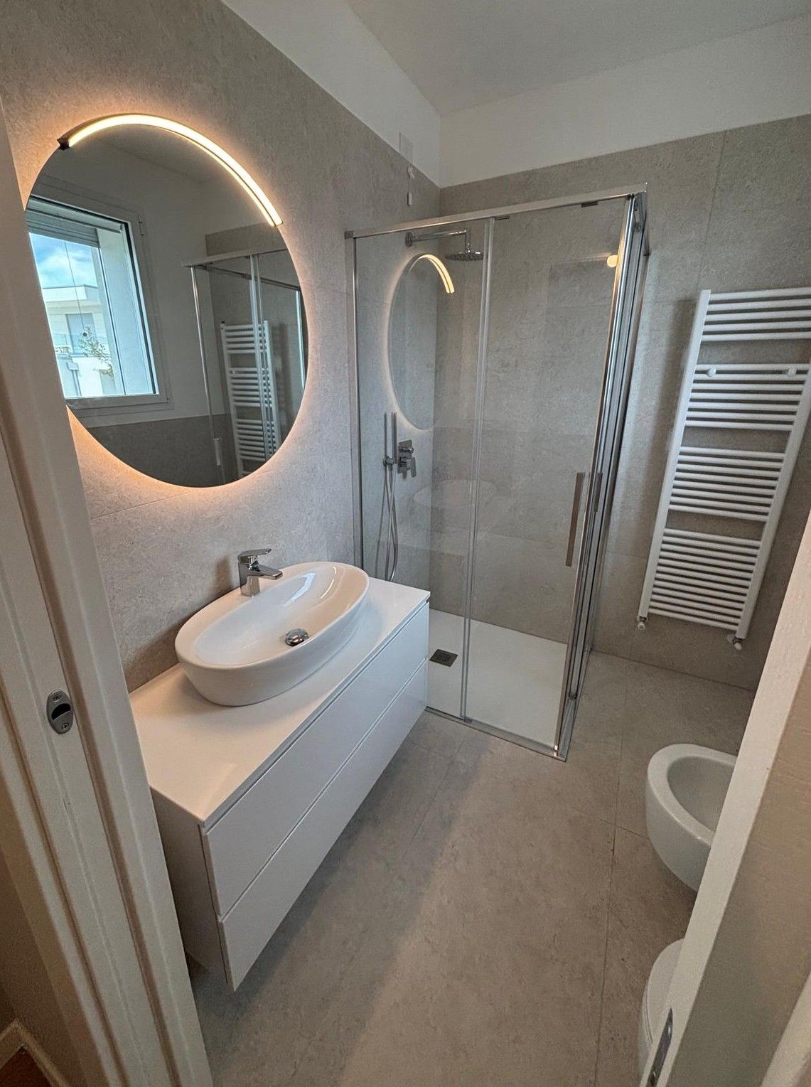 Bagno moderno con specchio rotondo illuminato, mobile lavabo bianco, doccia in vetro e termoarredo.