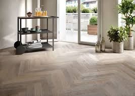 Soggiorno moderno con pavimento in parquet chiaro a spina di pesce, pianta e carrello bar accanto a una luminosa porta a vetri.