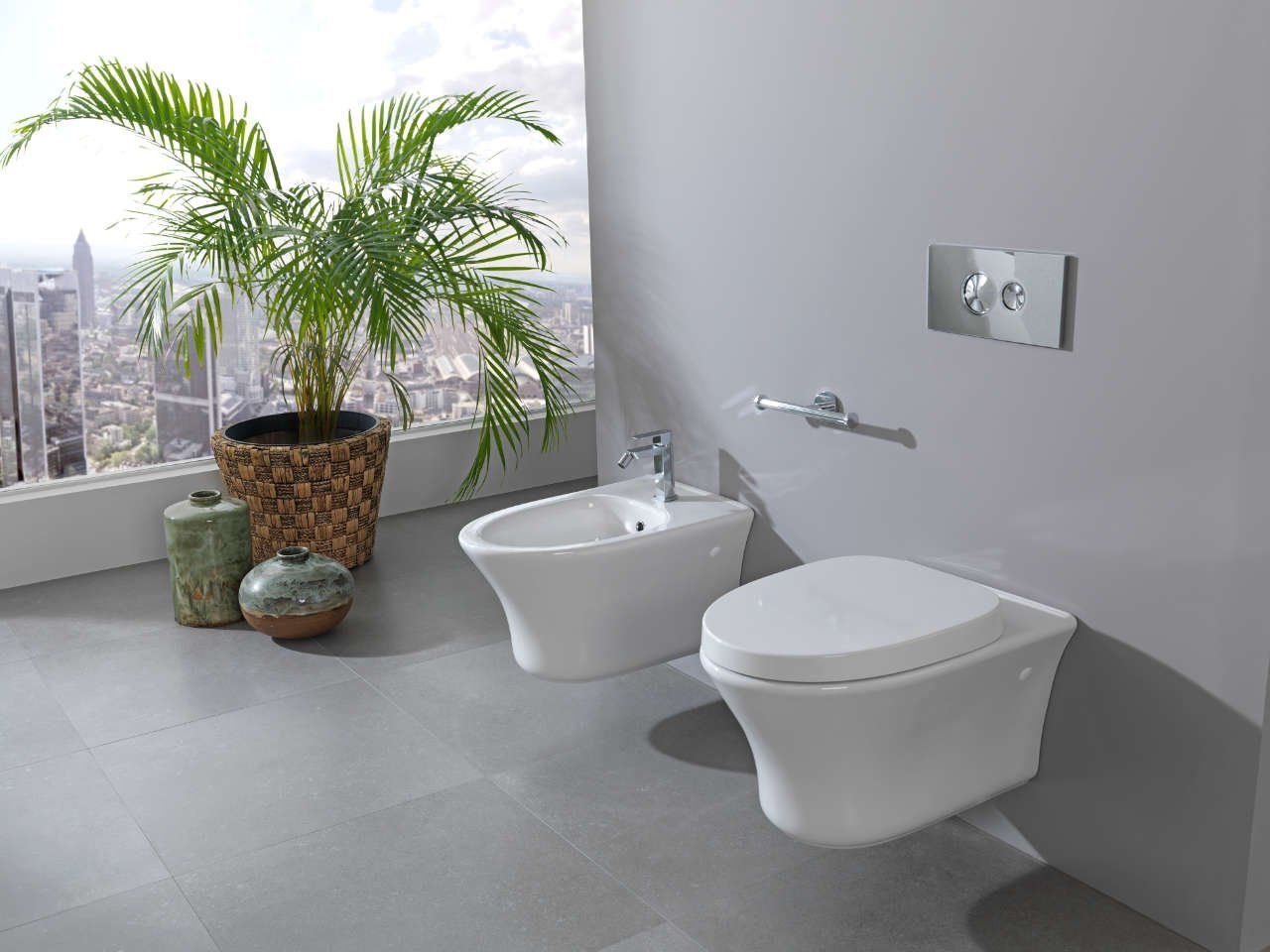 Bagno moderno con bidet e WC bianchi accanto a una palma in vaso vicino alla finestra