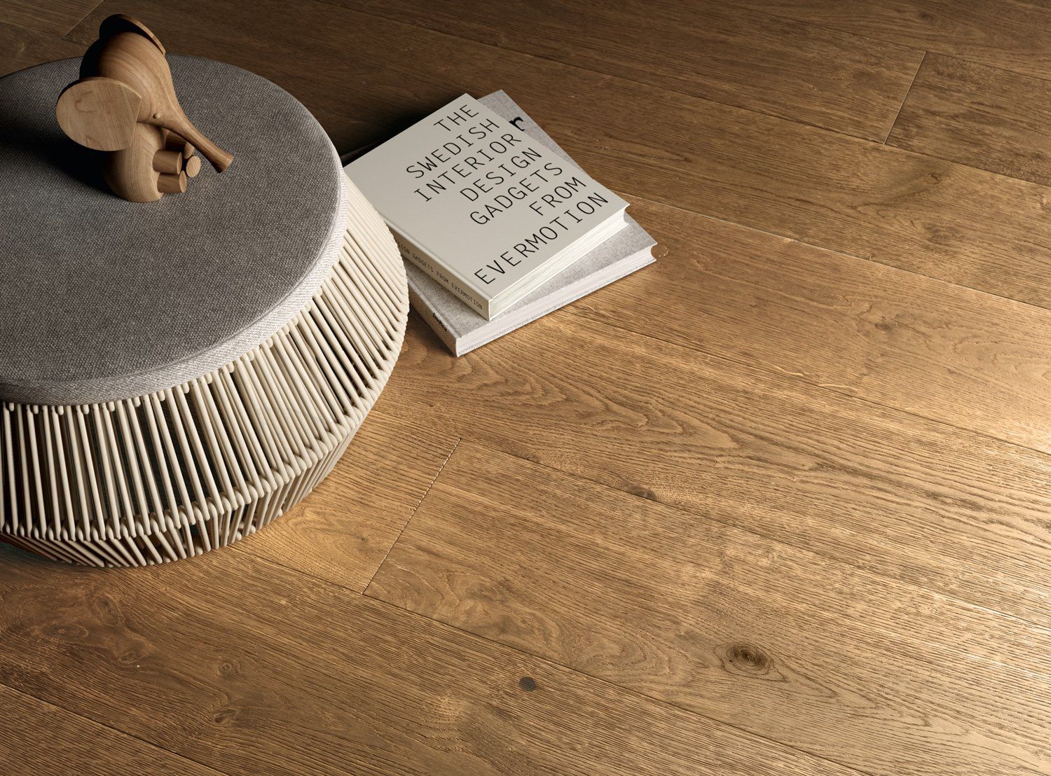 Pouf grigio intrecciato con un piccolo oggetto beige e un libro bianco su un pavimento in legno.