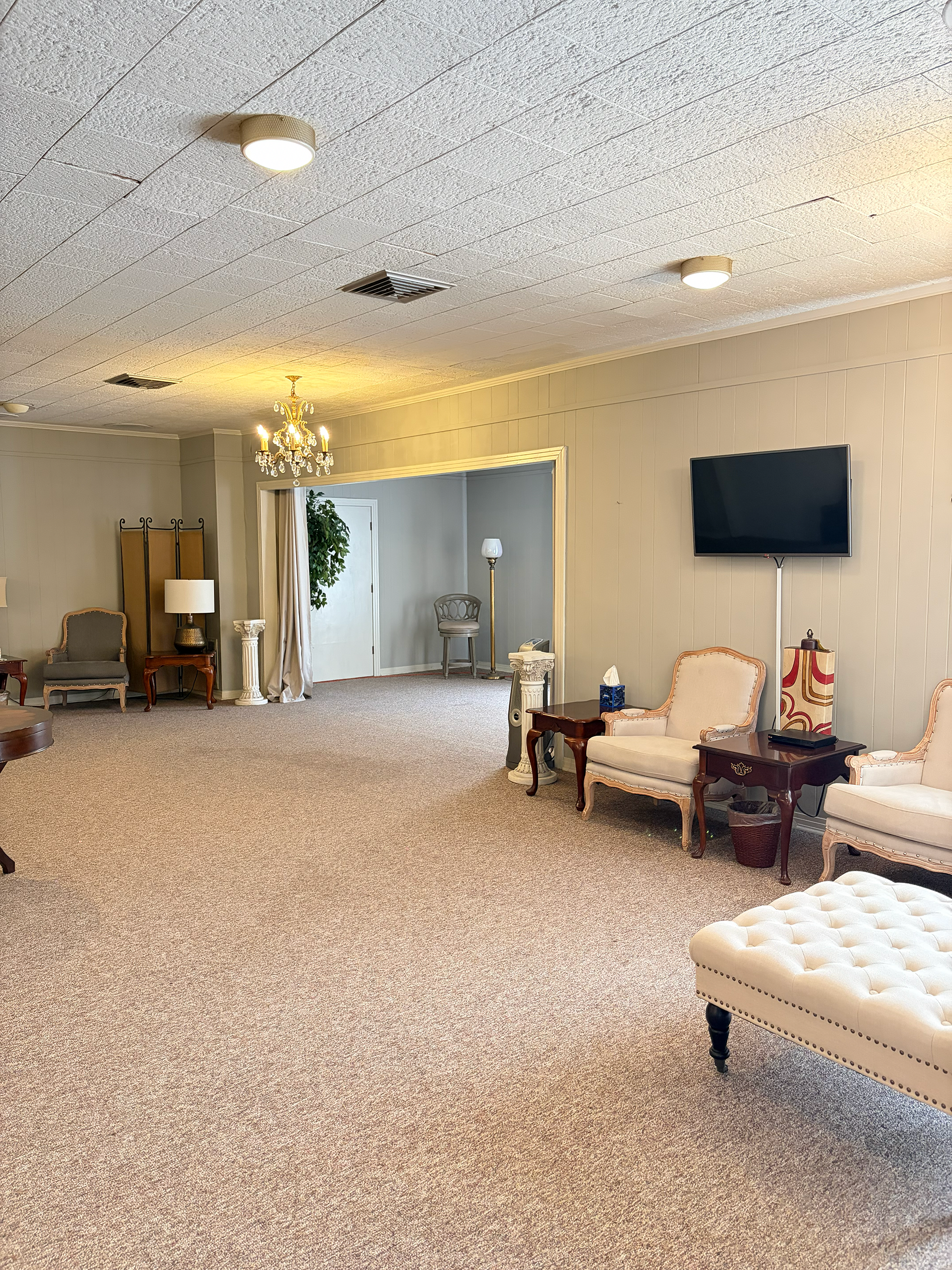 Spacious beige living room with carpet, wall TV, tufted bench, chairs, and open doorway to another room