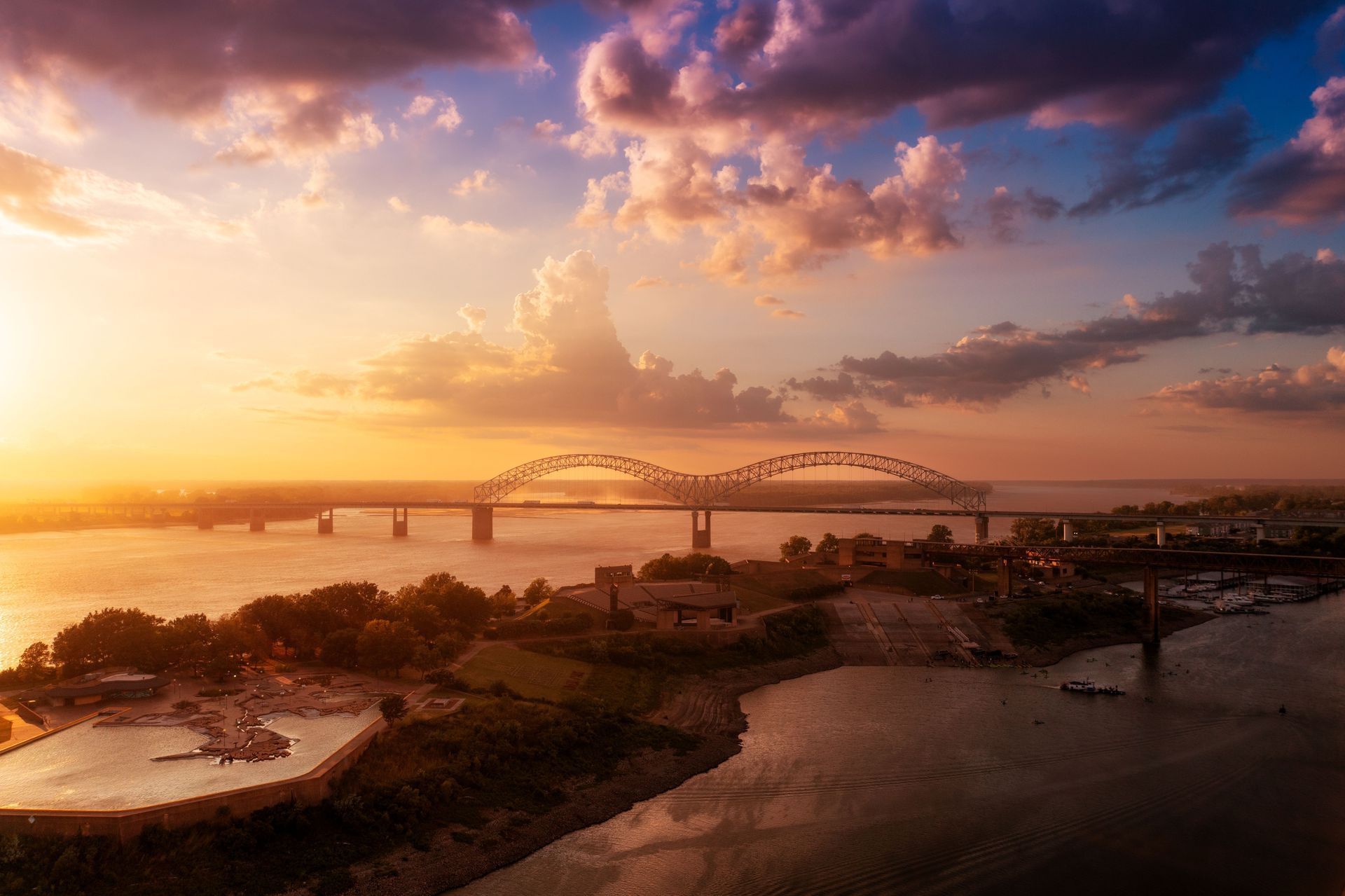 Sunset over a river with an arched bridge, city skyline, and glowing clouds