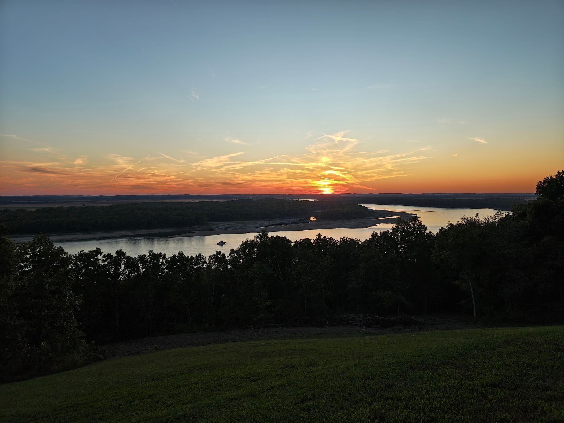 Sunset over a river with orange sky, silhouetted trees, and a grassy foreground.