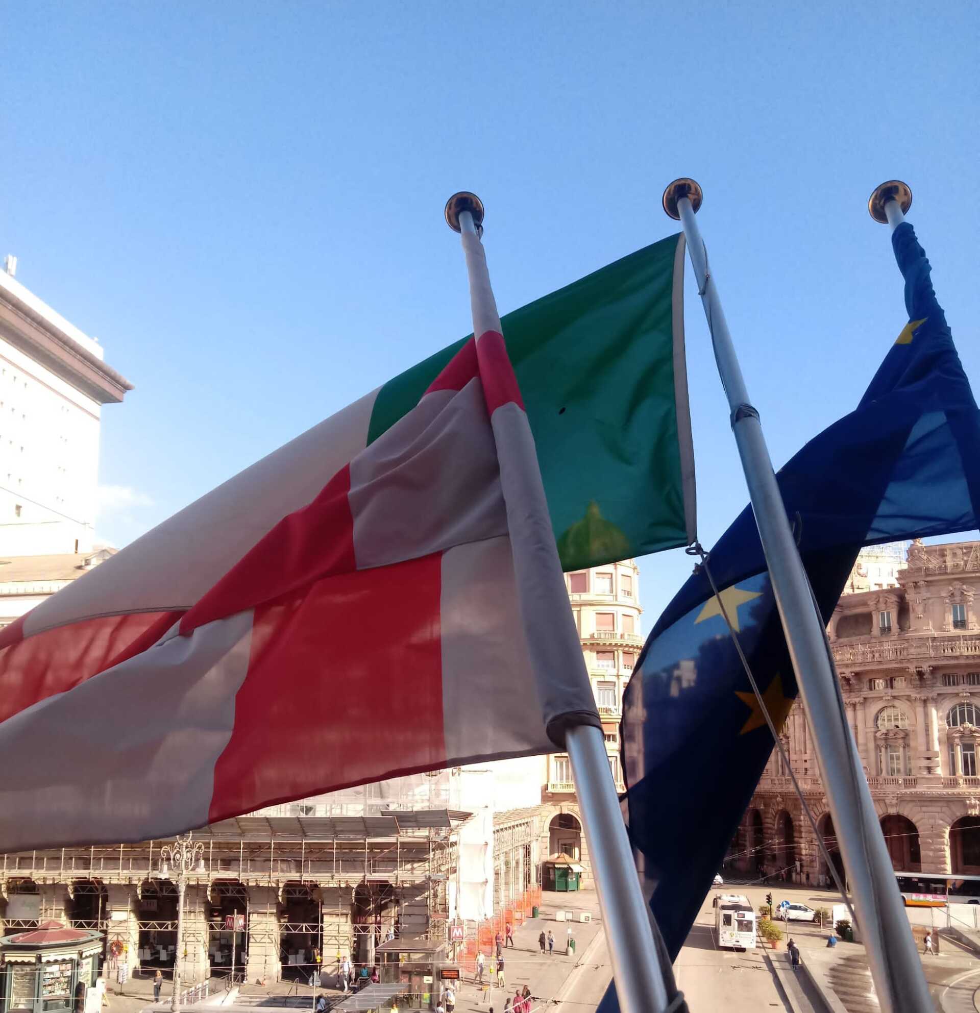 Bandiere italiana e dell'Unione Europea sventolano al vento. Sullo sfondo una piazza cittadina.