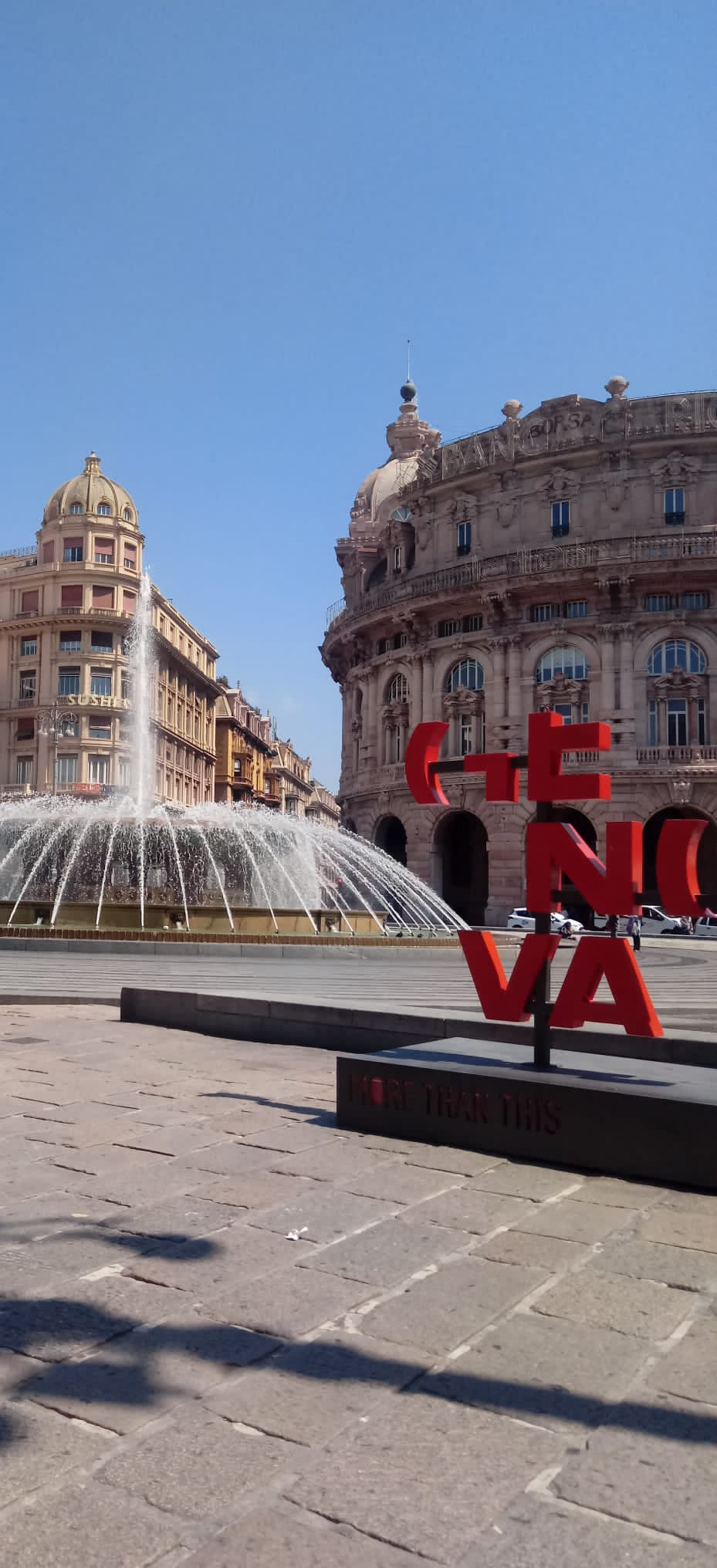 Una piazza pubblica di Genova, in Italia, con una grande fontana, edifici storici e un cartello rosso con la scritta