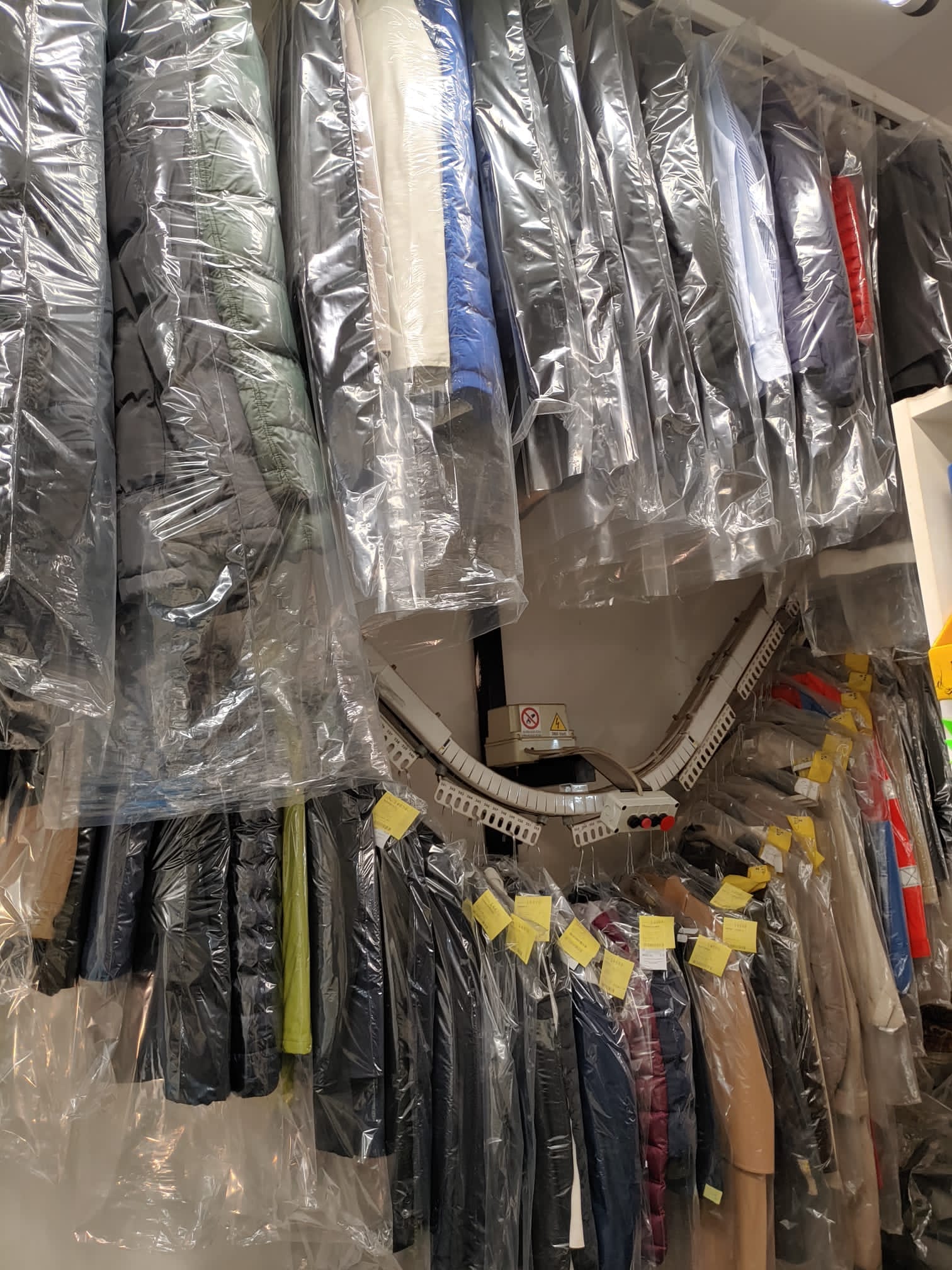 Cappotti avvolti in fodere di plastica appesi agli scaffali di una lavanderia a secco, per lo più di colore scuro.