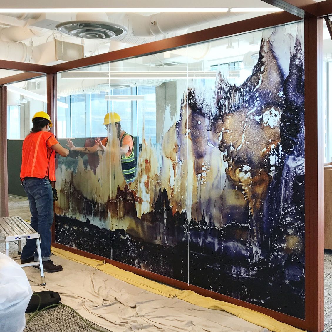 Two workers in yellow hard hats paint a large abstract mural on a glass wall indoors.
