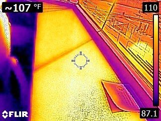 Thermal view of a sunlit walkway or deck beside a brick wall, with bright heat spots and a door mat.