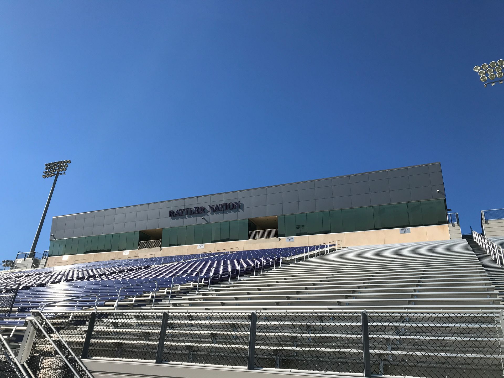 Empty stadium bleachers beneath a clear blue sky, with a large scoreboard and floodlights.