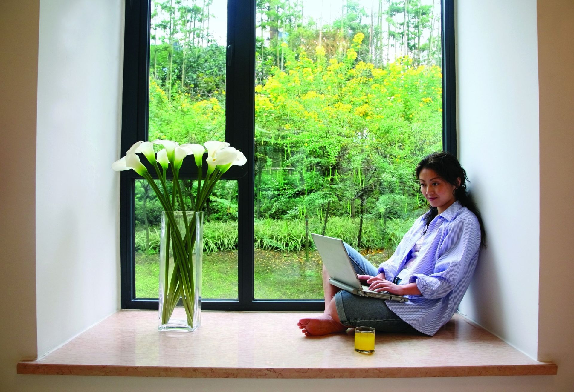 Person with laptop sitting by large window beside a vase of white flowers