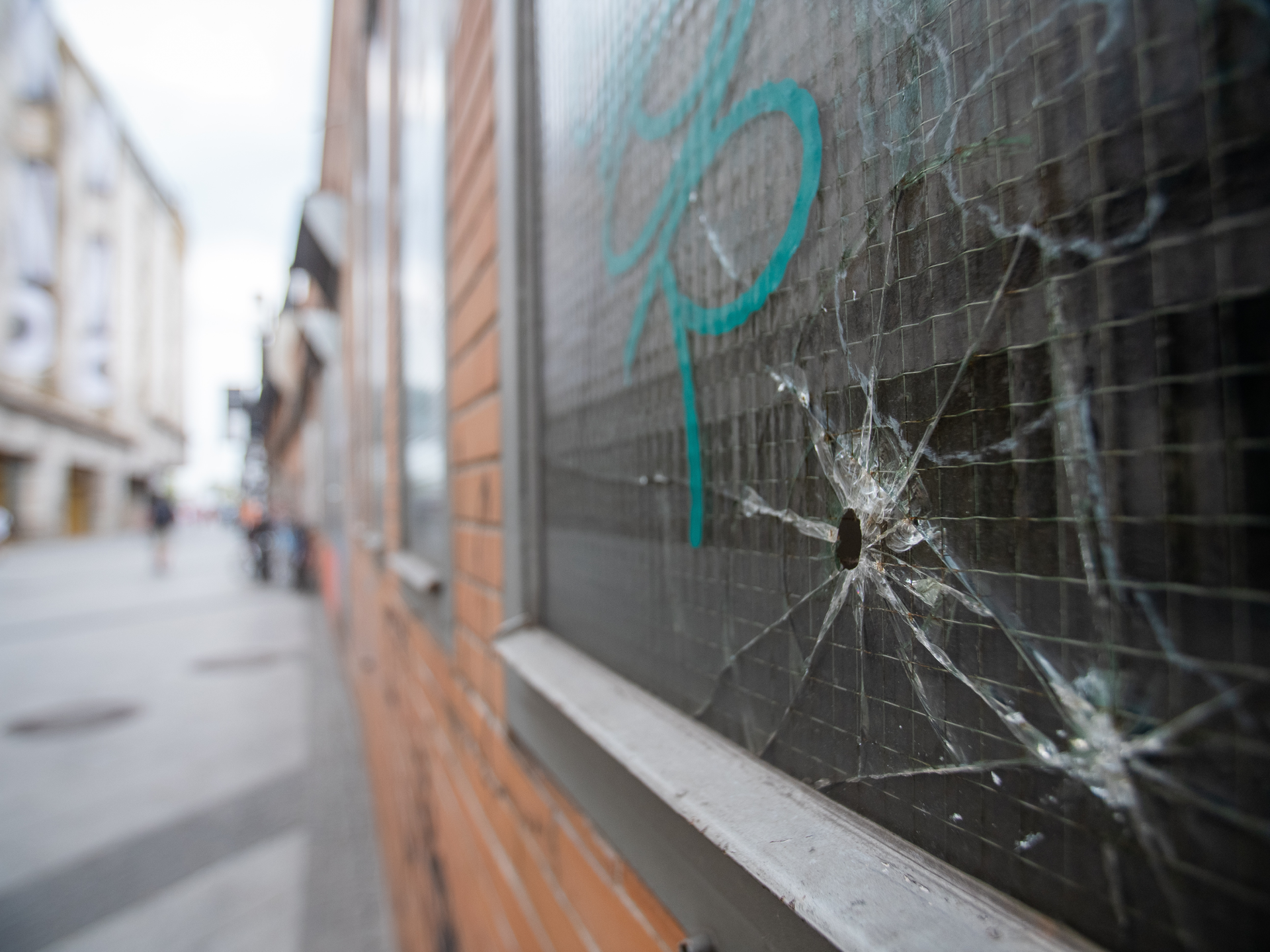 Cracked window beside a brick wall on a city sidewalk, with blurred buildings in the background