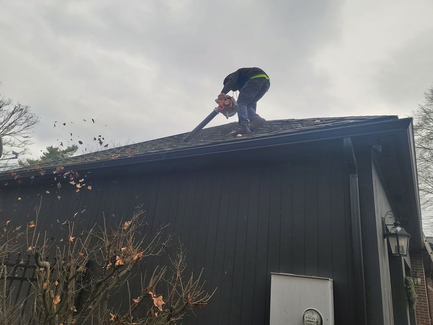 A person stands on the sloped roof of a dark building, using a leaf blower to clear debris from the shingles.