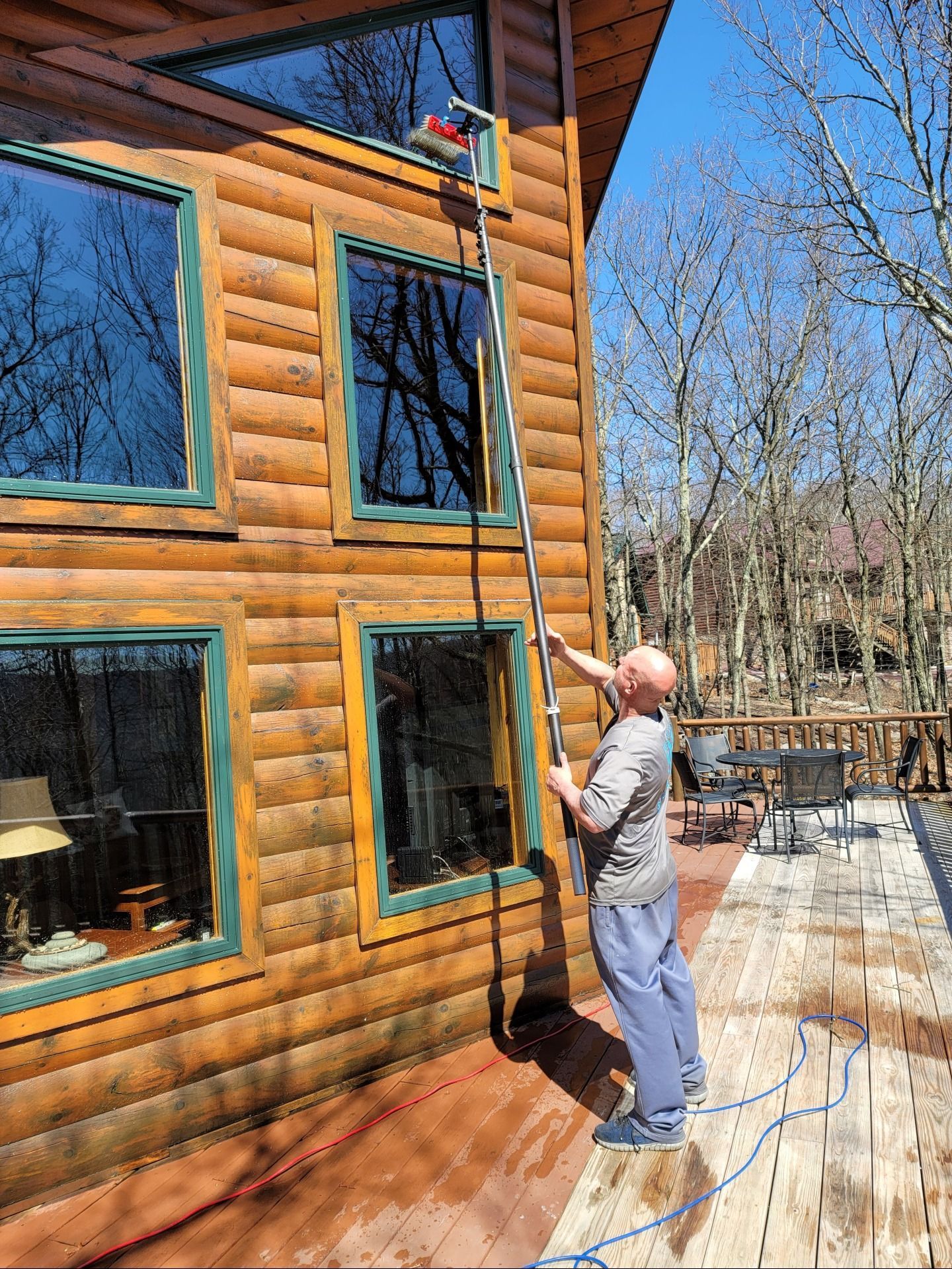 A person uses a long extendable pole with a brush to clean a high window on the side of a log cabin on a sunny day.