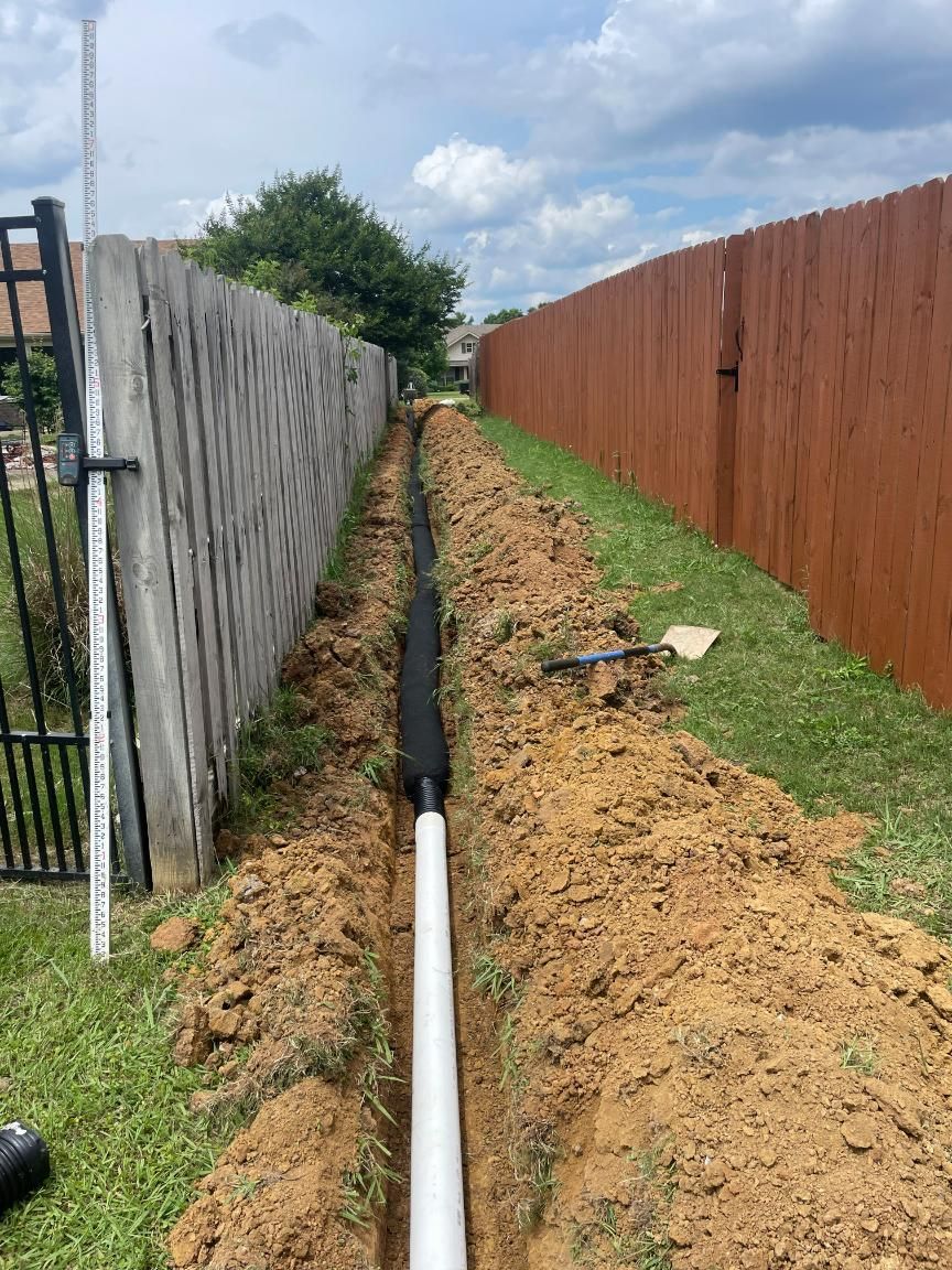 Trench with white pipe and black fabric next to a fence, on a sunny day.